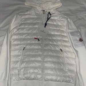 Vit moncler cardigan - Snygg vit pufferjacka från Moncler med quiltad framsida, huva och dragkedja. Jackan har ribbade muddar, två fickor med dragkedja och en patch med Moncler-logga på ärmen. Materialmix med polyester och bomull för en clean och fräsch look. Storleken är XL men den är liten i storlek så den sitter som en L 