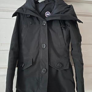 Svart vinterjacka från Canada Goose - Säljer en svart Canada Goose vinterjacka med huva och stora knappar framtill. Jackan har dragkedja på ärmen, två fickor med lock och klassisk Canada Goose-logga på axeln. Perfekt för kalla dagar och riktigt snygg streetstil. Jackan är också använd.