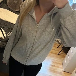 Grå hoodie med dragkedja Gina Tricot - Mysig grå hoodie från Gina Tricot i storlek S. Tröjan har huva med snörning, dragkedja framtill och långa ärmar. Perfekt för chill dagar eller när du vill ha en enkel och stilren look. Tillverkad i mjuk bomullsblandning.