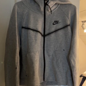 Grå Nike hoodie med dragkedja - Snygg grå hoodie från Nike med svart logga på bröstet och svarta detaljer. Tröjan har hel dragkedja framtill, huva  och två fickor. Perfekt för en sportig och avslappnad stil.
