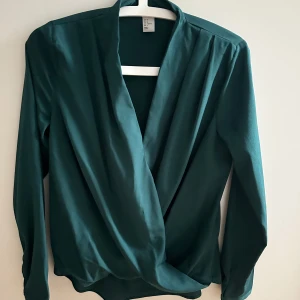 Grön omlottblus från H&M - Snygg grön omlottblus från H&M med långa ärmar och djup v-ringning. Blusen har en elegant omlottfront och är tillverkad i ett mjukt, slätt polyestertyg som faller fint. Perfekt att styla med jeans eller kjol för en trendig look. Tyvärr har blusen ett par defekter vid sidan
