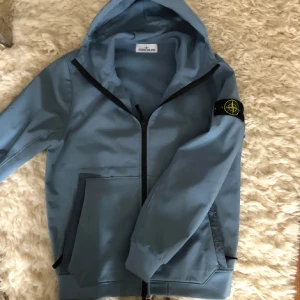 Blå softshelljacka från Stone Island - Snygg blå softshelljacka från Stone Island med huva och svart dragkedja. En perfekt höst och vinter jacka.
