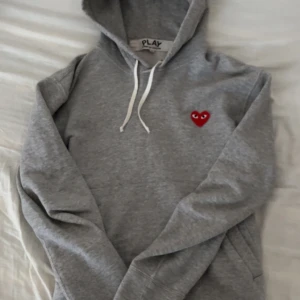 Grå hoodie Comme des Garçons Play S - Grå hoodie från Comme des Garçons Play i 100% bomull. Klassisk modell med huva, vita snören och det ikoniska röda hjärta-märket på bröstet. Långärmad och med känguruficka framtill. Perfekt för en chill och stilren look.