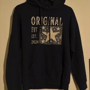 Mörkblå hoodie från Scndi  - Säljer en mörkblå hoodie från Scndi med tryck framtill som har stjärnor och texten 'ORIGINAL FIT EST. 2024'. Tröjan har huva med snörning och en stor magficka. Väldigt eftertraktad. Perfekt för dig som gillar en avslappnad och cool stil.