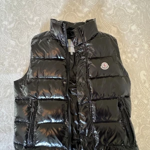 Svart dunväst från Moncler - Snygg svart dunväst från Moncler med glansig finish och klassisk logga på bröstet. Västen har hög krage, dragkedja och tryckknappar framtill samt två fickor med dragkedja. Perfekt för lager-på-lager och streetstyle.