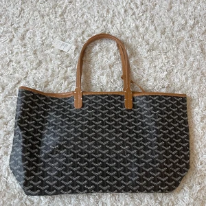 Svart shoppingväska med mönster - Säljer en ikonisk svart shoppingväska med klassiskt grafiskt mönster och bruna skinnhandtag. Väskan har ljus insida och medföljande liten clutch i samma stil. Perfekt för dig som vill ha en rymlig och snygg väska med lyxig känsla.