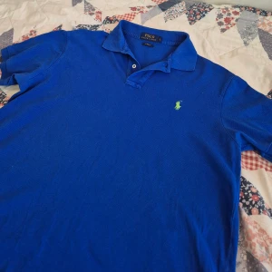Polo Ralph Lauren piké - Snygg blå piké från Polo Ralph Lauren i klassisk pikéstil med krage och knappslå. Custom fit-modell med korta ärmar och ikonisk grön broderad logga på bröstet. Perfekt för dig som gillar stilren och sportig vibe.