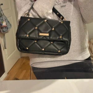 Svart quiltad handväska med guldnitar - Snygg svart handväska i quiltad design med guldfärgade nitar och detaljer. Väskan har ett handtag upptill och en stilren, lyxig look. Tillverkad i skinnimitation och perfekt för dig som gillar statement-accessoarer.