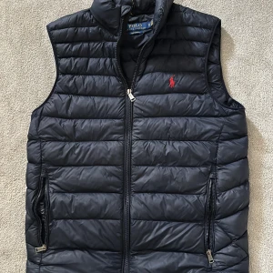 Svart dunväst från Polo Ralph Lauren - Snygg svart dunväst från Polo Ralph Lauren med horisontella quiltade paneler och klassisk ståkrage. Västen har dragkedja framtill, två sidofickor med dragkedja och röd broderad logga på bröstet. Perfekt för lager-på-lager och chill stil.