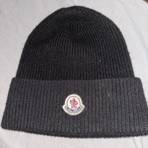 Svart Moncler mössa - Snygg svart ribbstickad mössa från Moncler med klassisk uppvikt kant och broderad logotyp framtill. Mössan är tillverkad i mjuk ull och har en stilren, enkel design som passar perfekt till kalla dagar. Loggan har detaljer i vitt, blått och rött.