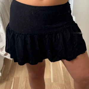 Svart volangkjol från NA-KD x Amela - Supersöt svart kortkjol från NA-KD x Amela med volangkant och elastisk midja. Kjolen är i mjuk bomull och har en flowy passform som ger en chill vibe. Perfekt att styla med crop top eller oversized hoodie.