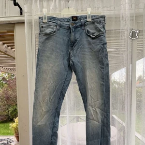 Blå Lee jeans med slitningar - Säljer ett par klassiska blå jeans från Lee med snygga slitningar och femficksmodell. Jeansen har normal passform och är tillverkade i mjukt denimtyg. Perfekta för dig som gillar en avslappnad och tidlös stil.