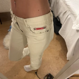 Vita randiga jeans från Blue Collection - Säljer ett par beige jeans från Blue Collection med diskreta vertikala ränder. Byxorna har klassisk femficksdesign, låg midja och raka ben. Snygga detaljer med röda logotaggar och dragkedja framtill. Perfekta för dig som gillar en clean och stilren look.