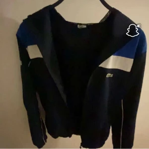 Blå och svart vindjacka från Lacoste - Snygg vindjacka från Lacoste i blått och svart med vita detaljer. Jackan har dragkedja framtill, meshfoder och klassisk Lacoste-logga på bröstet. Perfekt för dig som gillar sportig och stilren look.