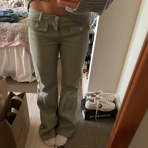 Olivgröna bootcut-byxor från Monki - Snygga olivgröna byxor från Monki med bootcut-snitt och hög midja. Byxorna har två stora fickor framtill med lock och knappar, samt bälteshällor. Tillverkade i mjuk bomull med lite stretch för extra komfort.