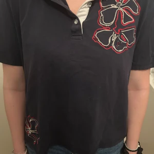 Mörkblå t-shirt med blommor från Basler - Mörkblå t-shirt från Basler med korta ärmar och krage. Framsidan har stora broderade blommor i vitt, rött och svart. Knappslå med två knappar framtill och kontrastfärgad insida vid halsen. Skön och klassisk modell i bomull.