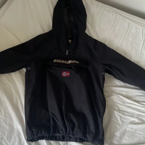 Svart vindjacka från Napapijri - Svart vindjacka från Napapijri med huva och norsk flagga på bröstet. Jackan har stor magficka. Perfekt för blåsiga dagar och har en clean, sportig vibe.