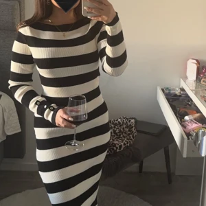 Randig långklänning i stickad bomull - Snygg långärmad långklänning med breda svarta och vita ränder. Klänningen är ribbstickad, har en tight passform och en slits på sidan. Ärmarna har dekorativa knappar vid handleden. Perfekt för dig som gillar en stilren och trendig look.