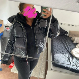 Svart pufferjacka från Tommy Hilfiger - Säljer en svart, glansig pufferjacka från Tommy Hilfiger med logga på ärmen. Jackan har dragkedja framtill, hög krage och avtagbar huva. Den är vadderad och har en oversized passform som ger en cool look. Perfekt för kalla dagar när du vill vara både varm och snygg.