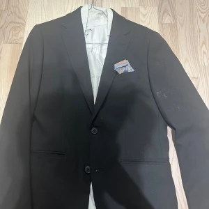 Svarta kostymbyxor från Riley  - Snygga svarta kostymbyxor med en klassisk design. Byxorna har en rak passform och är perfekta för formella tillfällen. De har en knapp och dragkedja framtill samt hällor för bälte.