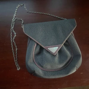 Yves Saint Laurent svart axelväska - Säljer en svart klassisk Vintage axelväska från Yves Saint Laurent med kedjerem och triangelformad silverdetalj (har lagt till kedjan själv)framtill. Väskan är i grovvävd textil med mörkröda skinnkanter och har innerfack med dragkedja. Perfekt för dig som gillar klassisk men edgy stil.