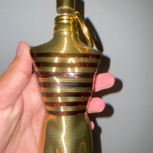 Jean Paul Gaultier Le Male Le Parfum - Säljer en ikonisk parfymflaska från Jean Paul Gaultier, Le Male Le Parfum. Flaskan är formad som en manlig torso i metallic guld med svarta och röda ränder och en guldfärgad ring vid halsen. Perfekt som statement på hyllan eller i samlingen.
