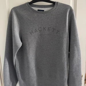 Hackett London tröja - Snygg grå sweatshirt från Hackett London med diskret logga framtill. Klassisk rund halsringning och långa ärmar. Mjuk och skön bomullsblandning som passar perfekt till jeans eller joggers. Enkel och stilren design för en avslappnad look.