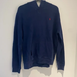 Mörkblå hoodie från Ralph Lauren - Säljer en mörkblå hoodie från Ralph Lauren med klassisk huva och känguruficka. Köp på KidsBrandStore. Tröjan har en liten röd broderad logga på bröstet och är tillverkad i mjuk bomull. Perfekt för en chill och avslappnad stil. Storlek s/170