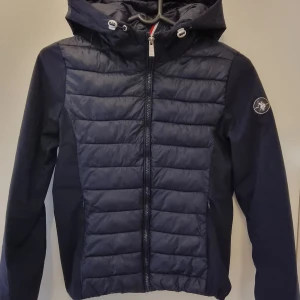 Mörkblå pufferjacka från U.S. Polo Assn. - Snygg mörkblå pufferjacka från U.S. Polo Assn. med huva och dragkedja framtill. Jackan har quiltad framsida, långärmade släta ärmar och logga på ena ärmen. Perfekt för kyliga dagar och riktigt stilren look. Står 38 i men liten så skulle säga 36. 