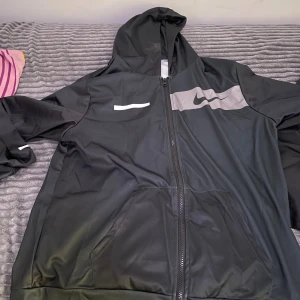 Svart vindjacka från Nike med huva - Säljer en svart vindjacka från Nike med huva och hel dragkedja. Jackan har en grå panel med Nike-logga på bröstet och praktiska fickor framtill. Insidan är ljusgrå och materialet känns lätt och smidigt, perfekt för blåsiga dagar.