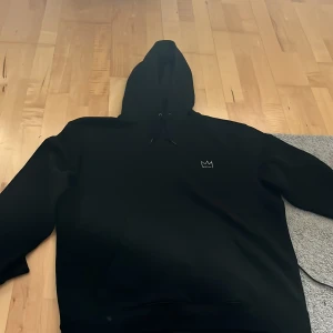Svart Hov1 hoodie XL Vi Dansar Vidare - Svart hoodie från Hov1 i storlek XL med broderad krona på bröstet och stor turnétext 'Vi Dansar Vidare I Livet' på ryggen. Klassisk huva med snörning och mjukt material, perfekt för dig som gillar streetwear och vill visa support för Hov1.