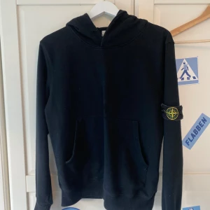 Svart hoodie från Stone Island - Svart hoodie från Stone Island med klassisk patch på vänster ärm. Tröjan har huva, känguruficka och långa ärmar. Använt den fåtal gånger och har inga stora skavanker eller hål eller fläck. Vid frågor eller nåt är de bara att höra av er! Tröjan sitter som en S