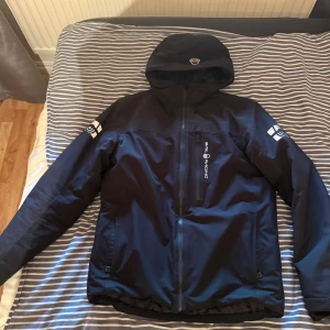 Svart vinter jacka från Sail Racing - Säljer en svart vinter jacka från Sail Racing med huva och snygga loggor på ärmarna. Jackan har dragkedja framtill och en bröstficka med Sail Racing-tryck. Perfekt för blåsiga dagar och stilren look.