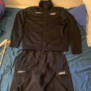 Svart Nike tracksuit i polyester - Säljer en svart tracksuit/tränings overall från Nike i storlek L. Tröjan har dragkedja framtill, höga muddar och sportiga vita tryck på bröstet. Materialet är mjukt och stretchigt polyester, perfekt för träning eller chill. Klassisk Nike-logga och modern look.