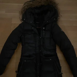 Hollies Vinterjacka - Säljer en svart lång pufferjacka från Hollies med stor huva och äkta päls runt kanten. Jackan har flera fickor framtill, dragkedja och knappar. Perfekt för kalla dagar och riktigt snygg siluett. Huvan är avtagbar och jackan är quiltad för extra värme. (Priset kan diskuteras)