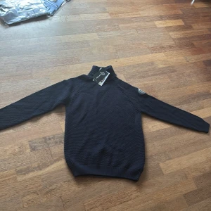 Mörkblå stickad polotröja Pelle P - Snygg mörkblå stickad polotröja från Pelle P med hög krage och diskret logomärke på ärmen. Tröjan har grovstickad struktur, ribbade muddar och är perfekt för kyliga dagar. Klassisk och stilren design som funkar till det mesta.