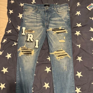 Blå ripped jeans från Amiri med patchar - Säljer ett par blå jeans från Amiri med grymma slitningar och stora bokstavspatchar i svart och vitt på både fram- och baksidan. Jeansen har klassisk femficksdesign, skinny passform och coola detaljer som trasiga partier med svarta ränder under. Perfekt för dig som gillar streetstyle. Byten av andra jeans kan även funka det med