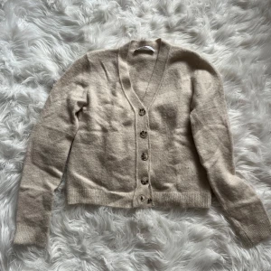 Beige kofta från Zara i ullmix - Mysig beige kofta från Zara i storlek M, tillverkad i mjuk ullmix. Koftan har v-ringad hals, ribbade muddar och fem knappar framtill. Perfekt för lager-på-lager och chill dagar. Enkel och stilren design som funkar till allt.