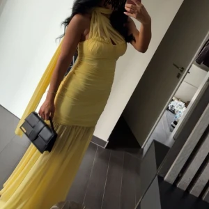 Gul långklänning med omlott och slits - Supersnygg gul långklänning med omlott-detalj vid halsen och en cool slits nedtill. Klänningen har figurnära passform, draperad mesh och axelbandslös design. Perfekt för dig som vill sticka ut med en ljus och fräsch look. Materialet är lätt och luftigt.