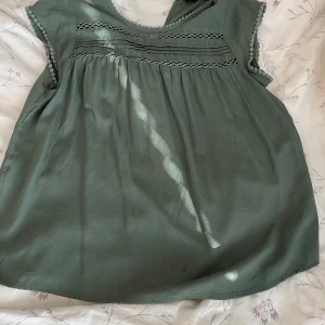 Grön blus från Vero Moda - Supersöt grön blus från Vero Moda med spetsdetaljer och små bollar längs ärmslut. Blusen har lös passform, rund halsringning och är ärmlös. Perfekt för dig som gillar en chill och luftig stil.