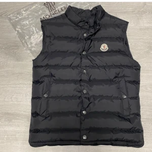 Moncler  - Snygg svart dunväst från Moncler med klassisk logga på bröstet. Västen har hög krage, knappar framtill och två fickor med lock. Perfekt för lager-på-lager och ger en clean, sportig look.