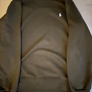 Svart sweatshirt från Polo Ralph Lauren - Svart sweatshirt från Polo Ralph Lauren i storlek M/L. Klassisk design med rund hals, långa ärmar och liten vit broderad logga på bröstet. Mjuk och skön bomullskvalitet, perfekt för en clean och stilren look.