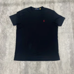 Svart t-shirt från Polo Ralph Lauren - Svart t-shirt från Polo Ralph Lauren med klassisk röd broderad logga på bröstet. Modellen är enkel med rund hals och korta ärmar. Tillverkad i mjuk bomull som känns skön mot huden. Perfekt för dig som gillar stilrena och ikoniska plagg.