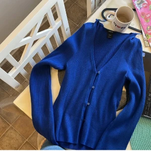 Kornblå ribbad kofta från Lindex - Kornblå ribbstickad kofta från Lindex i storlek M. Koftan har v-ringning, långa ärmar och knäppning framtill med matchande blå knappar. Perfekt att slänga över en t-shirt eller linne för en chill och snygg look.