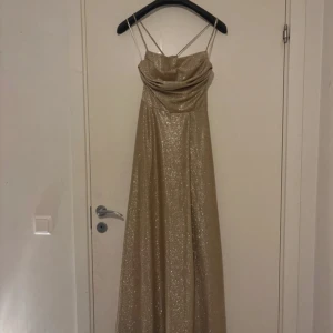 Guldig glittrig bal/bröllopsklänning  - Supersnygg långklänning i guld med glittrigt tyg och tunna axelband. Klänningen har snörning och öppen rygg, vilket ger. Perfekt för dig som vill sticka ut och glänsa på bal eller fest. och klänningen har en figurnära passform upptill.den köptes förra veckan när jag hade ett bröllop att gå på och är använd en gång så den är helt ny ungefär den är verkligen jätte fin men har tyvärr ingen användning till den länge och den köptes för 2500 från L&s i mobilia i Malmö 