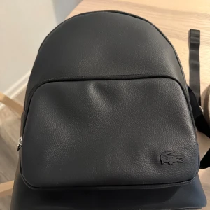 Svart ryggsäck från Lacoste i skinn - Snygg svart ryggsäck från Lacoste i stilren design med präglat skinn och klassisk krokodil-logga framtill. Väskan har justerbara axelband, mesh på baksidan för extra komfort och flera dragkedjefack. Perfekt för dig som vill ha en clean och modern look. Använd endast en gång 