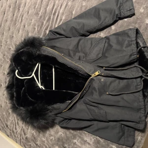 Svart parkajacka med fuskpäls - Säljer en svart parkajacka med mjukt fuskpälsfoder och stor huva med pälsdetalj. Jackan har dragkedja, snörning i midjan och stora fickor framtill. Perfekt för kalla dagar och riktigt snygg till streetwear-stilen.