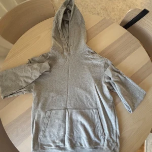 Grå hoodie från J.Lindeberg - Säljer en enkel grå hoodie med stor huva och magficka. Tröjan har långa ärmar och är gjord i mjuk bomull, perfekt för chill dagar. Diskret broderad text på bröstet. Passar dig som gillar klassisk streetstyle.