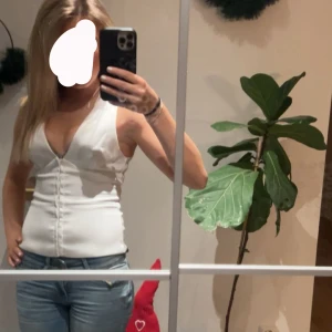 Vit v-ringad ärmlös topp - Säljer en stilren vit ärmlös topp med djup v-ringning och knapprad framtill. Sitter tight och har en clean look som passar perfekt till jeans eller kjol. Enkel att matcha och superfräsch till sommaren!