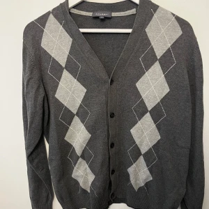 Grå kofta med argylemönster från Atlant - Snygg grå kofta från Atlant i bomull med klassiskt argylemönster i ljusgrått och vitt framtill. Koftan har v-ringning, knäppning med svarta knappar och ribbade muddar. Perfekt för lager-på-lager och en preppy look.